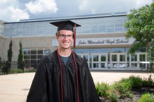 A graduate in cap and gown stands smiling in front of a modern school building on a sunny day.