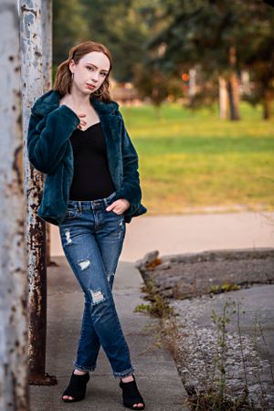 Model in distressed jeans and blue fur jacket poses casually against concrete pole in outdoor park setting.