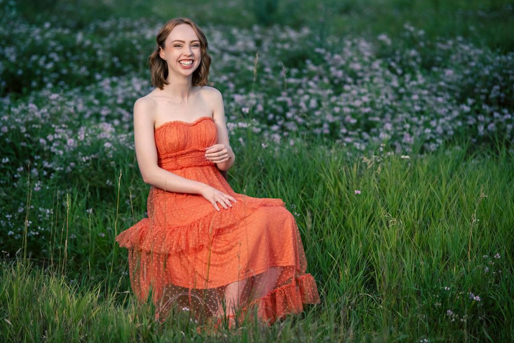 Someone in a coral-colored ruffle dress sits in a meadow of purple wildflowers on a summer evening.