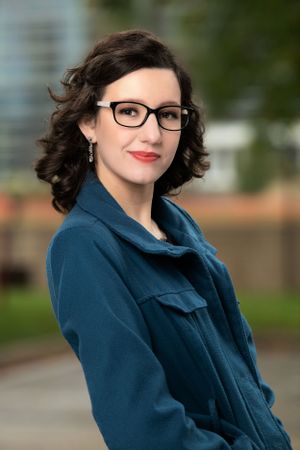 Professional portrait of someone wearing a navy blue coat and glasses with styled dark curly hair against a blurred outdoor background.