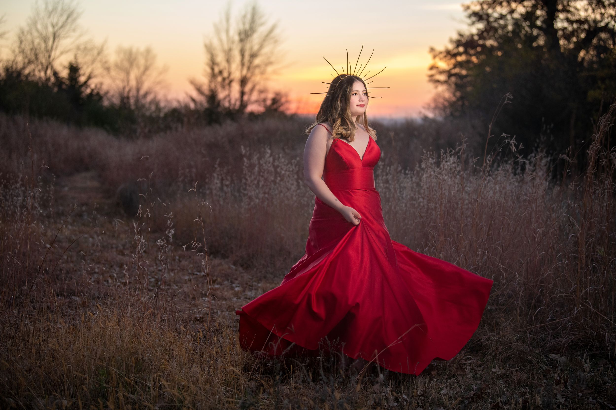 A flowing red gown twirls gracefully in a golden sunset field with bare trees silhouetted in the background.