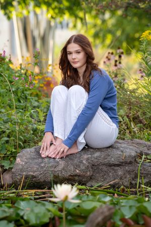 Someone in a blue sweater and white pants sits peacefully on a rock surrounded by flowers and greenery in a garden.