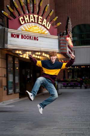 A person jumps joyfully in front of the illuminated Chateau Theater marquee at dusk, wearing a yellow and maroon striped sweater.