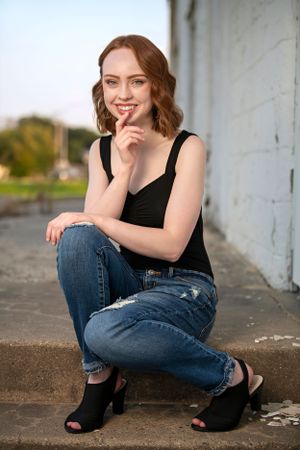 A woman with wavy hair poses outdoors wearing a black tank top and distressed jeans while sitting on concrete steps.