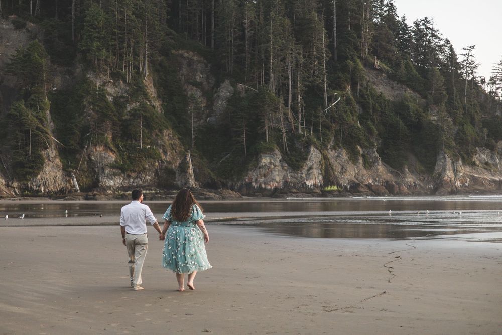 People walking along a scenic Pacific Northwest beach with towering cliffs and evergreen trees in the background.