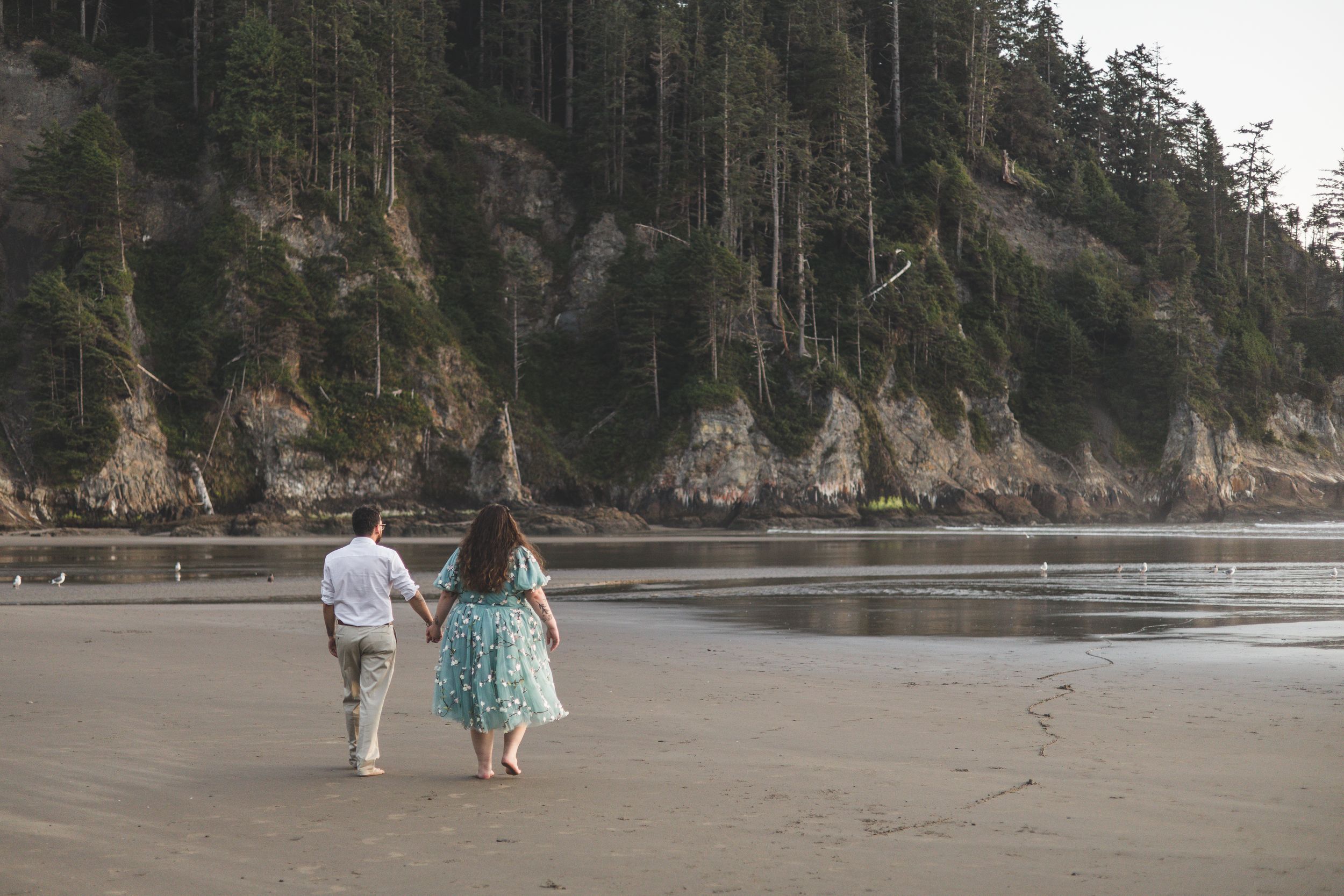 People walking along a scenic Pacific Northwest beach with towering cliffs and evergreen trees in the background.