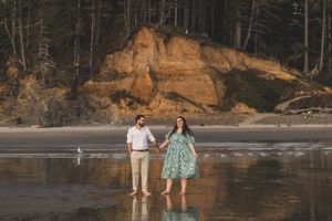 A couple walks along the shoreline of a scenic beach with tall cliffs and forest in the background.