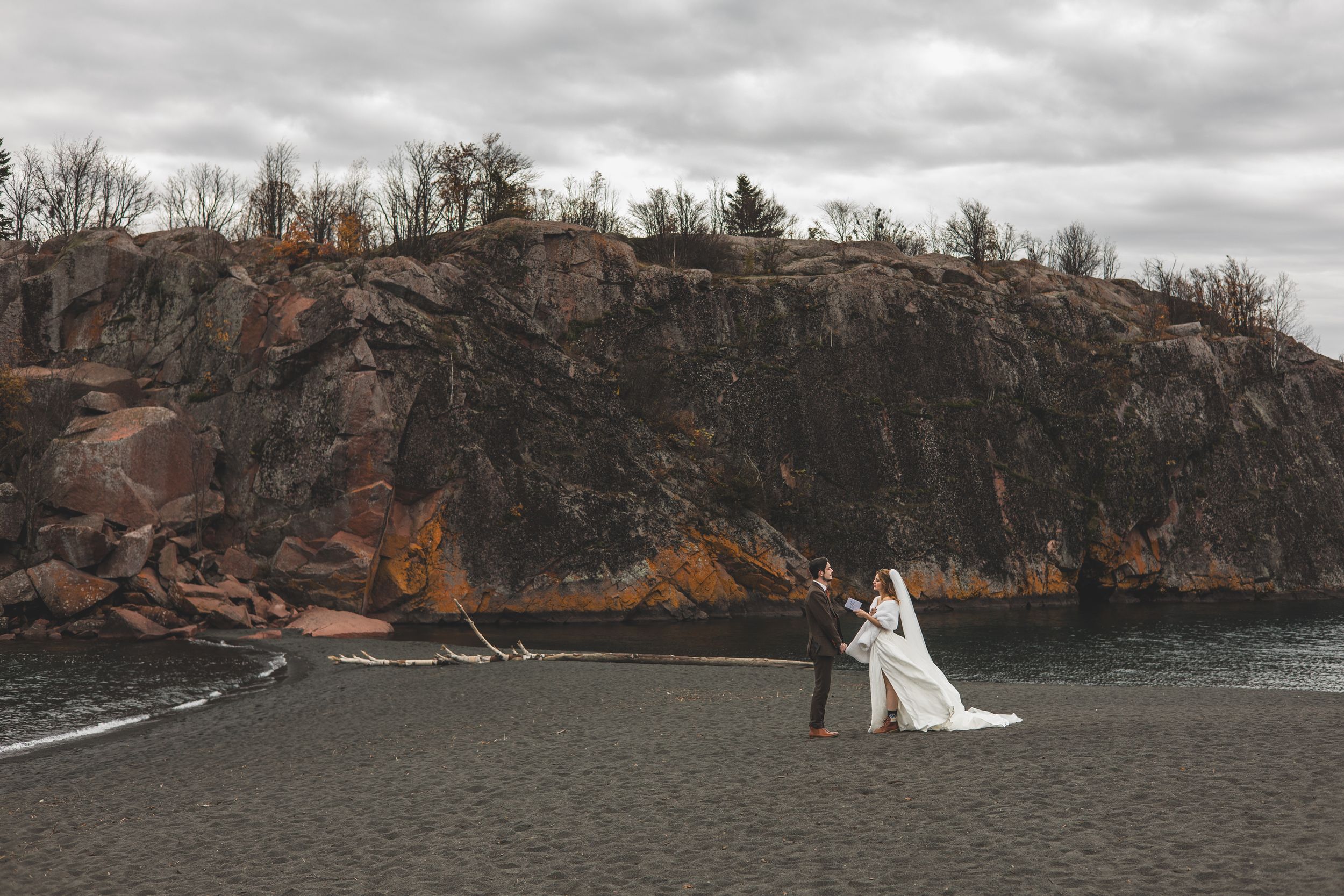 A wedding couple stands on a rocky cliff overlooking a dramatic lake landscape on an overcast day.