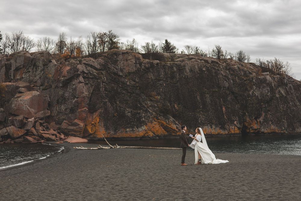 A wedding couple stands on a rocky cliff overlooking a dramatic lake landscape on an overcast day.