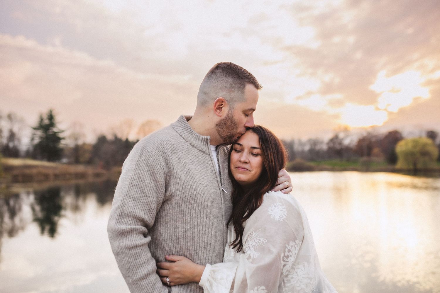 A couple embraces by a serene lake at sunset with autumn trees reflected in the calm water.