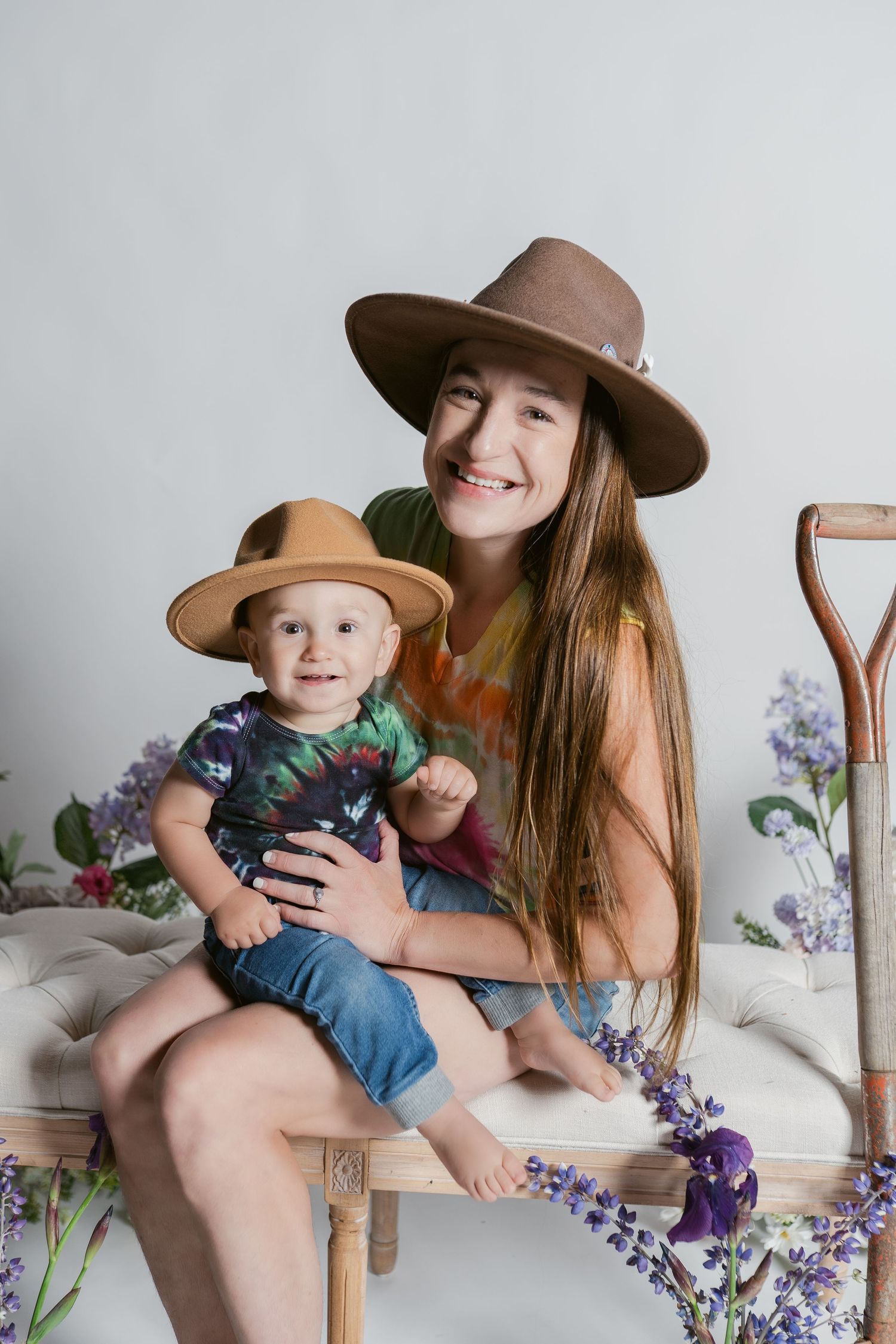 Mother and child wearing matching brown hats sit together on wooden chair surrounded by purple flowers.