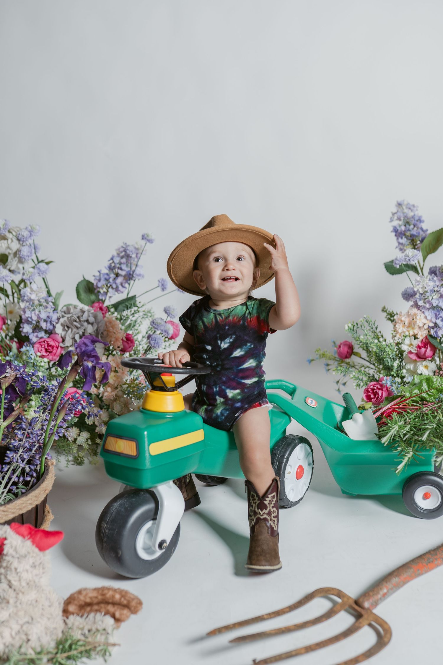 A young child wearing a brown hat and boots rides a green toy tractor amid colorful garden flowers and gardening tools.
