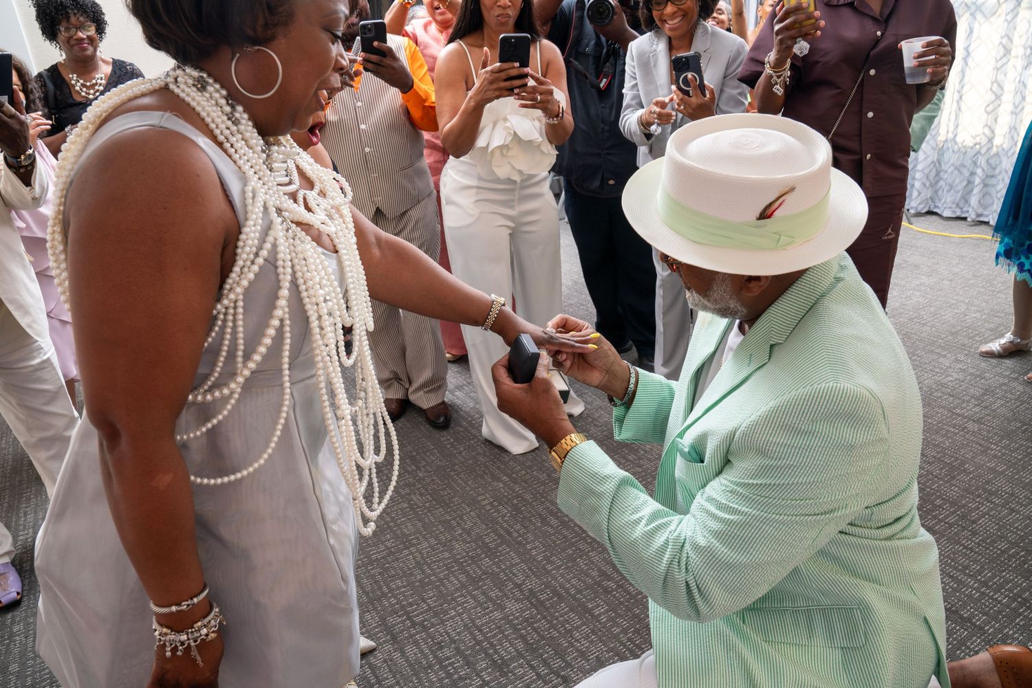 A person in a mint green suit and hat holds the hand of someone wearing a white dress and pearls at a social gathering.