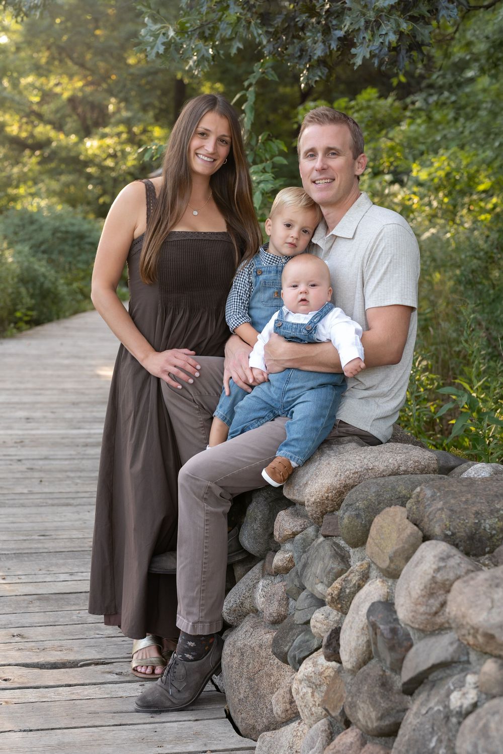 A smiling family of four with two young children posing outdoors on a wooden boardwalk beside a stone wall surrounded by green trees.