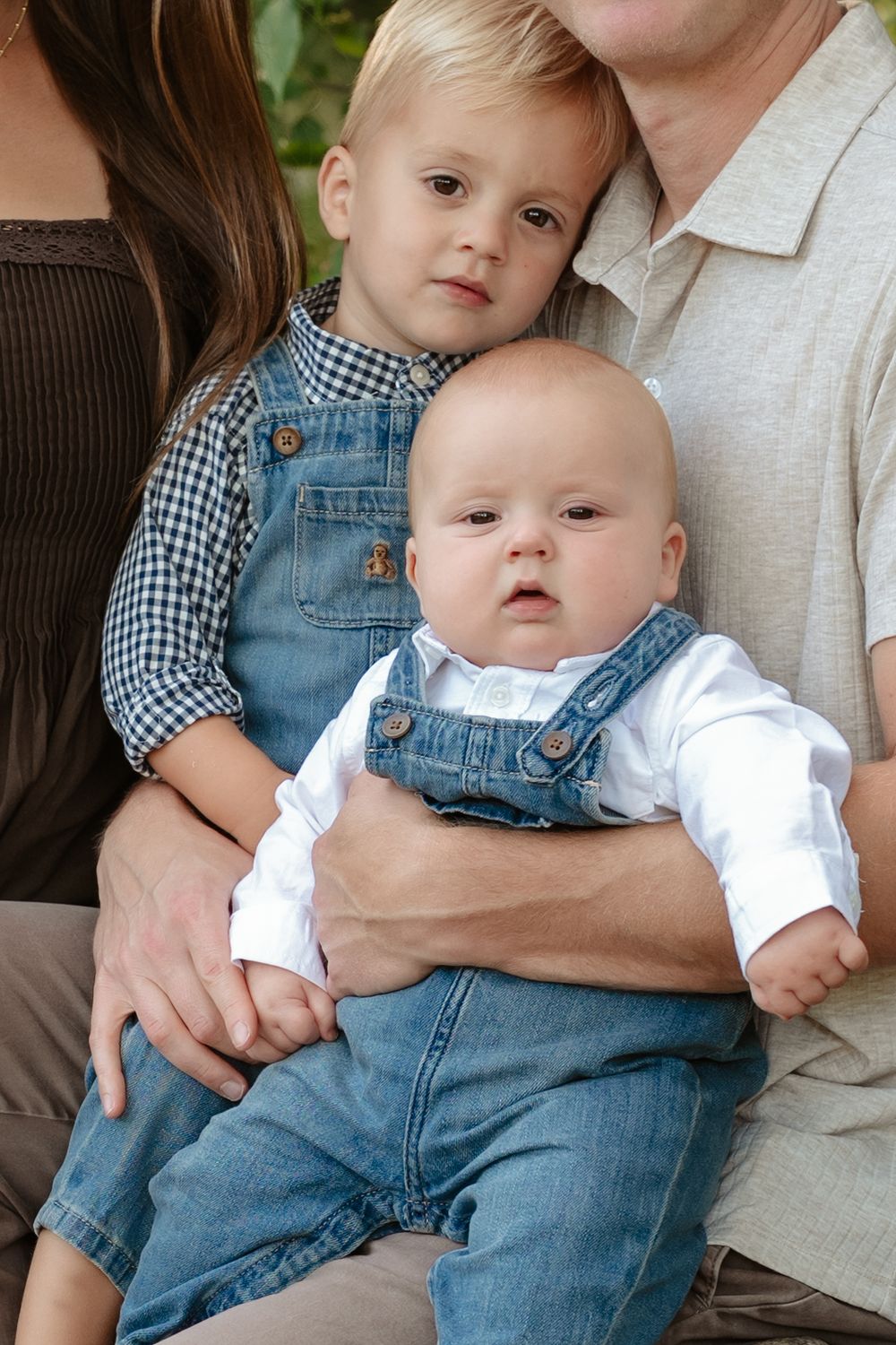Toddler and baby in matching denim overalls sit on the lap of a smiling adult during an outdoor family portrait.