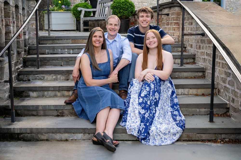 Family of four smiling together on stone steps outdoors, dressed in blue and white casual attire.