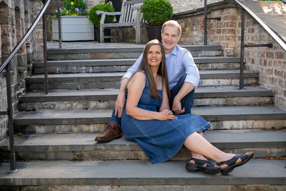 A smiling couple sits together on outdoor stone steps, woman in blue dress, man in light blue shirt.