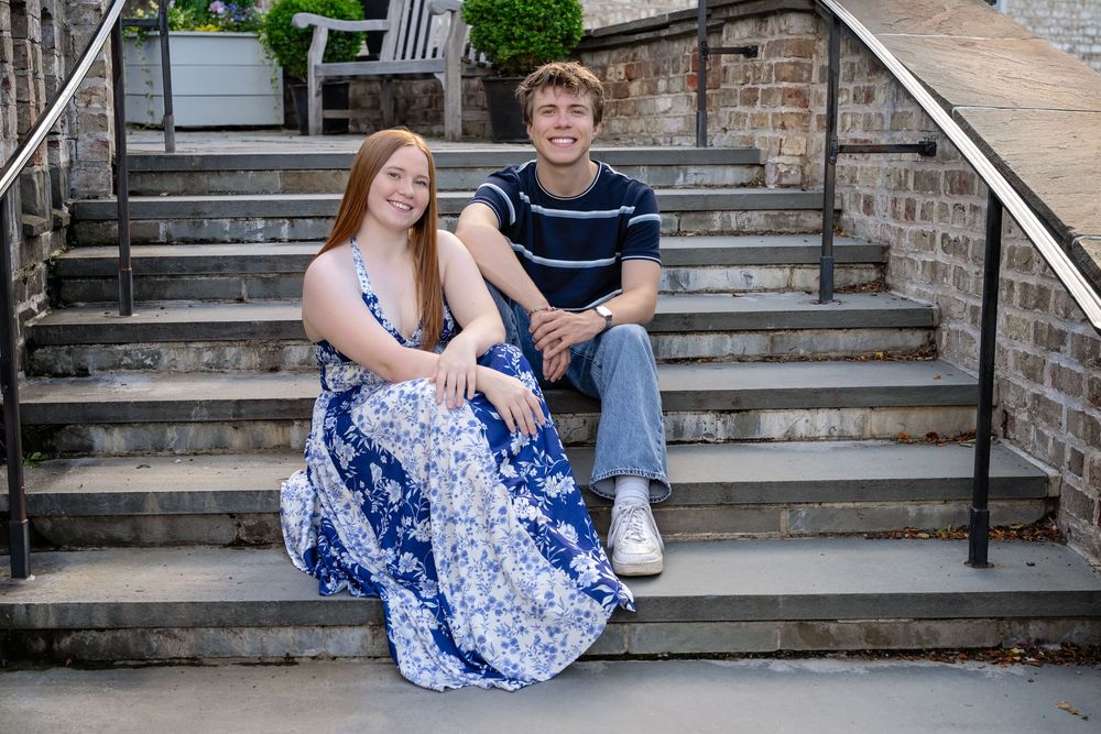 A young couple smiles while sitting on outdoor stone steps, the woman in a blue floral dress and the man in a striped shirt.