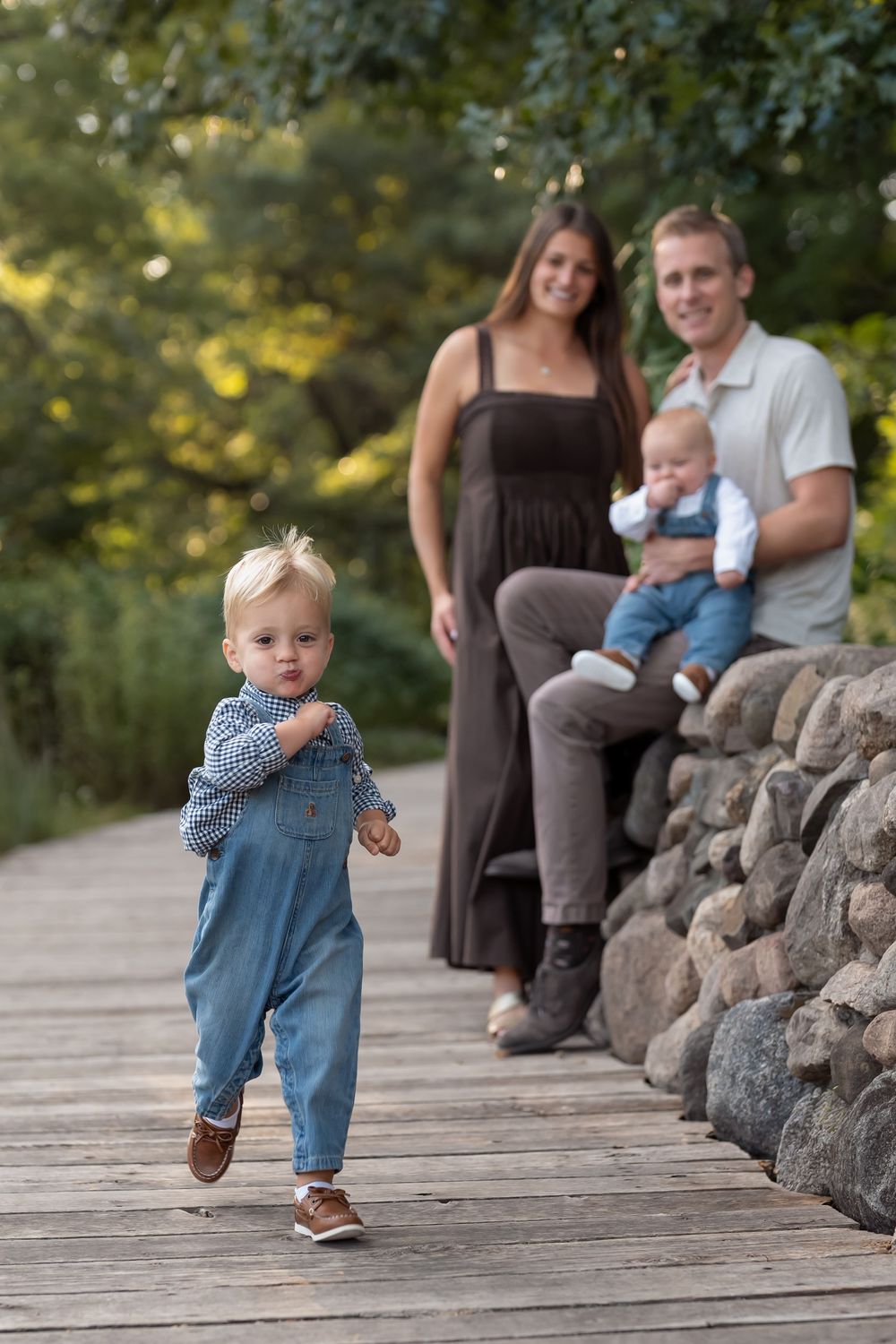 A smiling couple sits on a stone wall holding a baby while a toddler in denim overalls walks along a wooden boardwalk in a park.