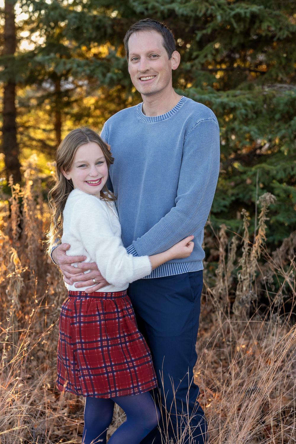 A man and young girl smile together outdoors among pine trees, she wears a plaid skirt and white sweater.