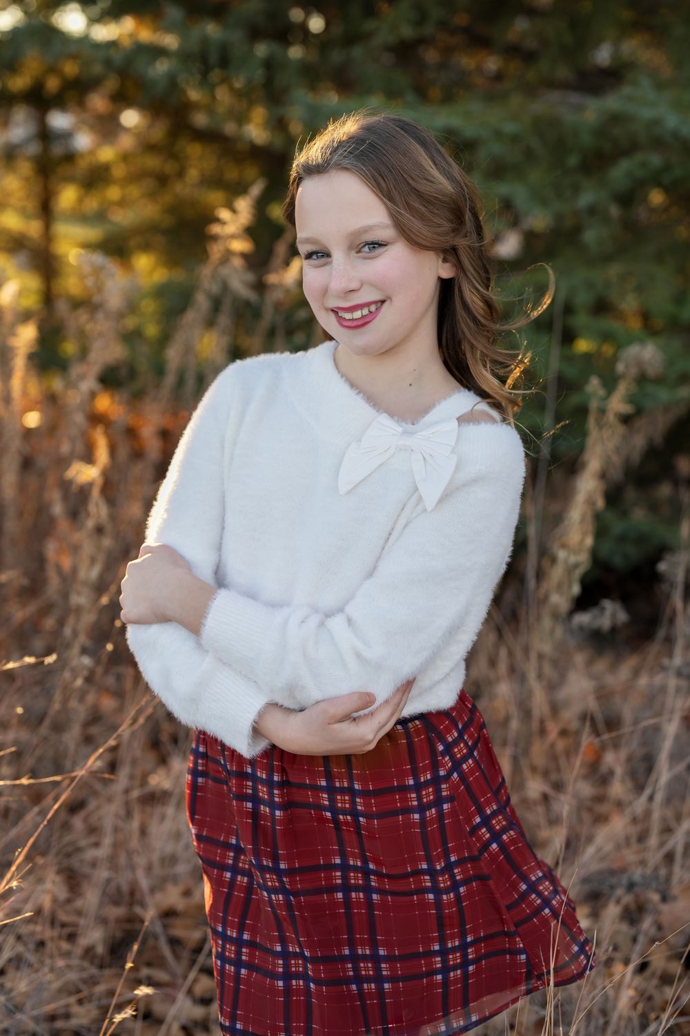 Smiling teenage girl in white fuzzy sweater with bow and red plaid skirt posing outdoors among dry grass and pine trees.
