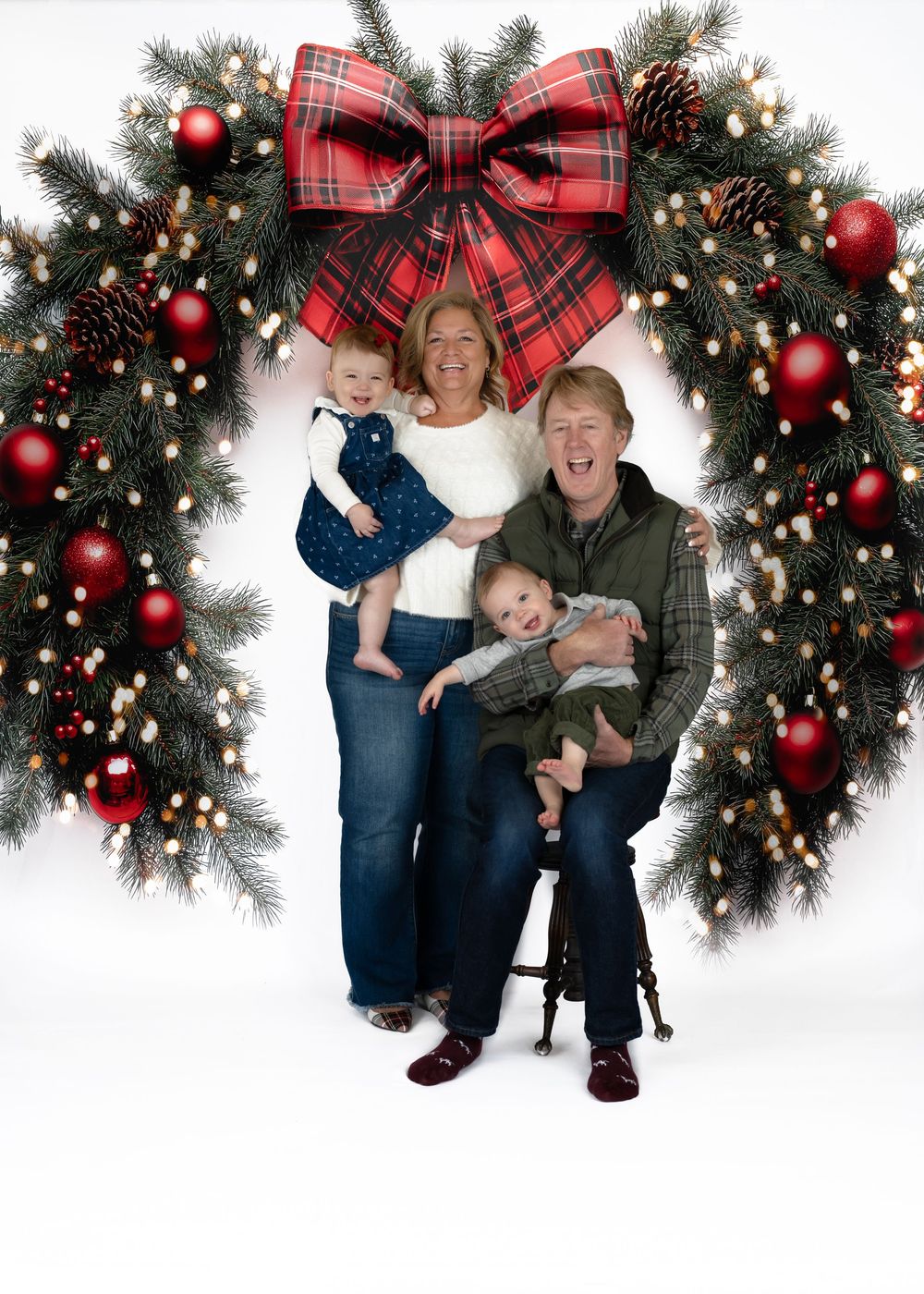 Smiling couple holds two young babies in front of a large decorated Christmas wreath in studio setting.