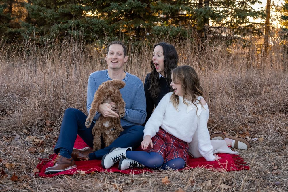 Family of three laughing outdoors on red blanket with brown puppy, surrounded by golden grass and pine trees.