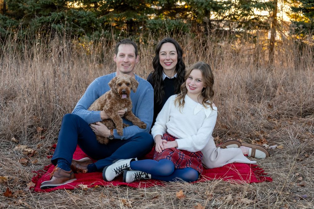 Family of two adults and a girl sit on red blanket outdoors with fluffy dog, smiling in a natural setting.