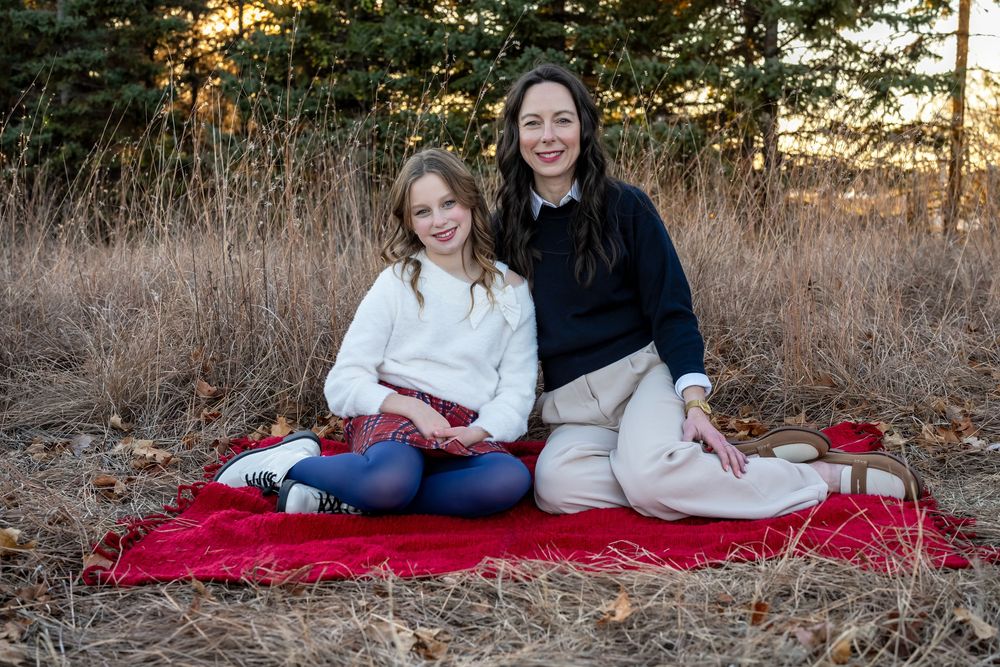 Woman and young girl smiling together on red blanket outdoors in autumn field setting.
