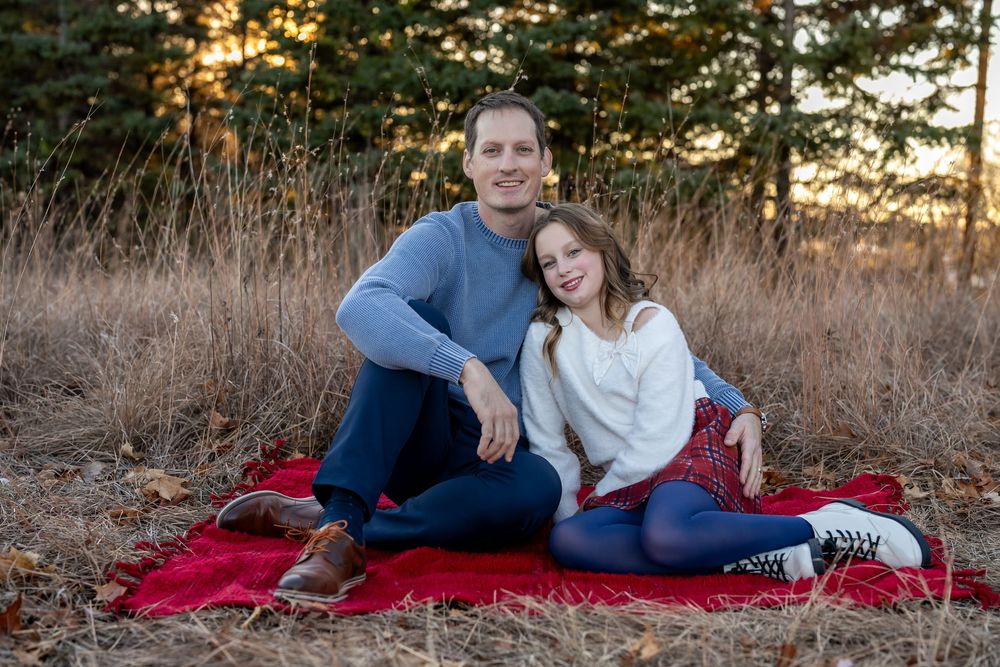 A man and young girl sit together on a red blanket outdoors, smiling warmly in a sunlit forest setting.