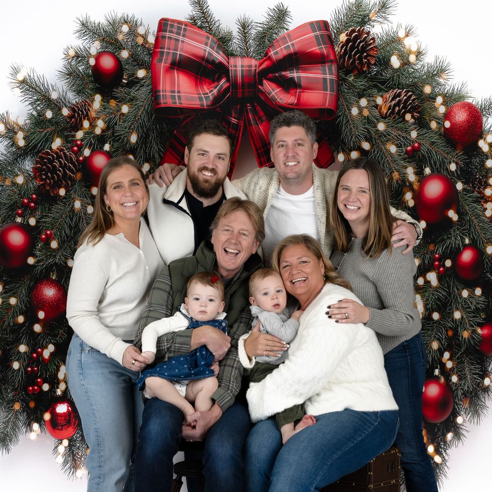 Happy multigenerational family with two infants smiles together in front of a decorated holiday Christmas wreath.