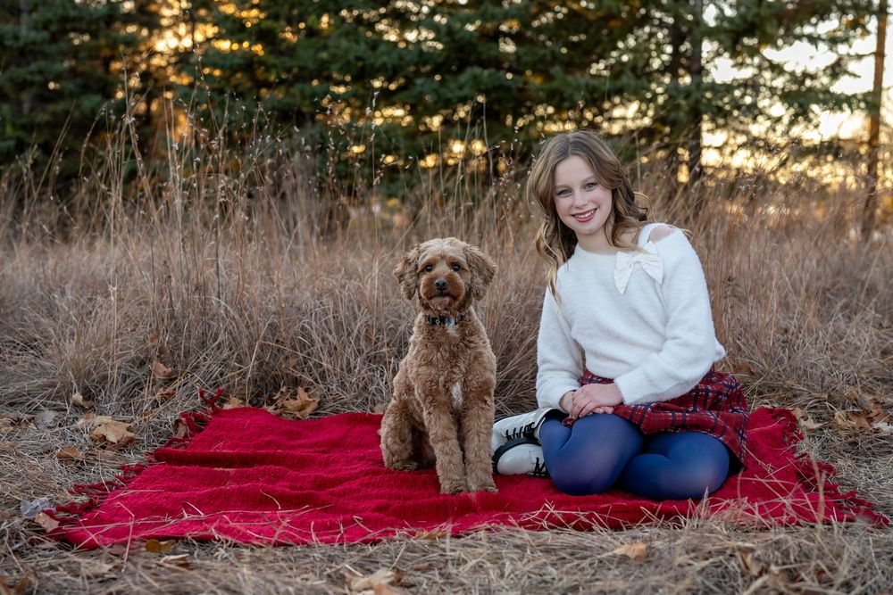Girl and brown doodle dog sitting on red blanket in autumn field with pine trees at golden hour.