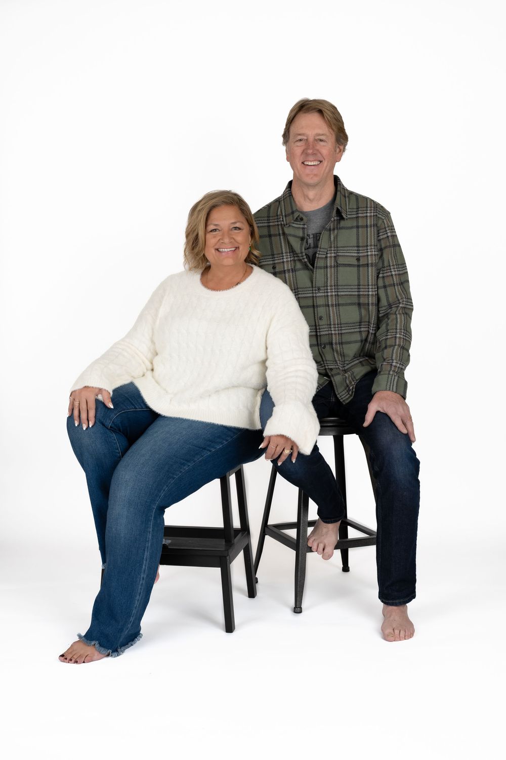 Smiling couple in casual attire sits on black stools barefoot against a clean white studio background.