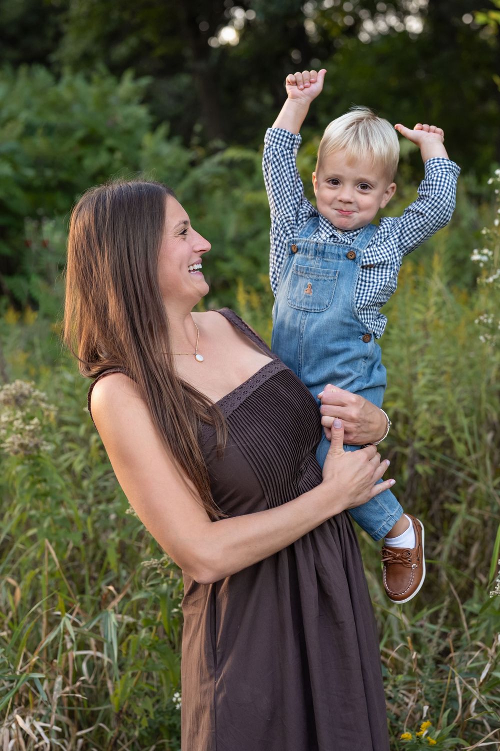 Smiling woman in a brown dress lifts a laughing toddler in denim overalls and a plaid shirt among tall green grasses outdoors.