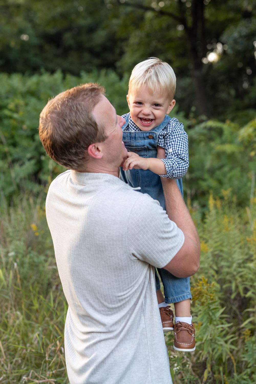 Smiling toddler in denim overalls and plaid shirt is held up by a man in a gray t-shirt in a sunny grassy meadow.