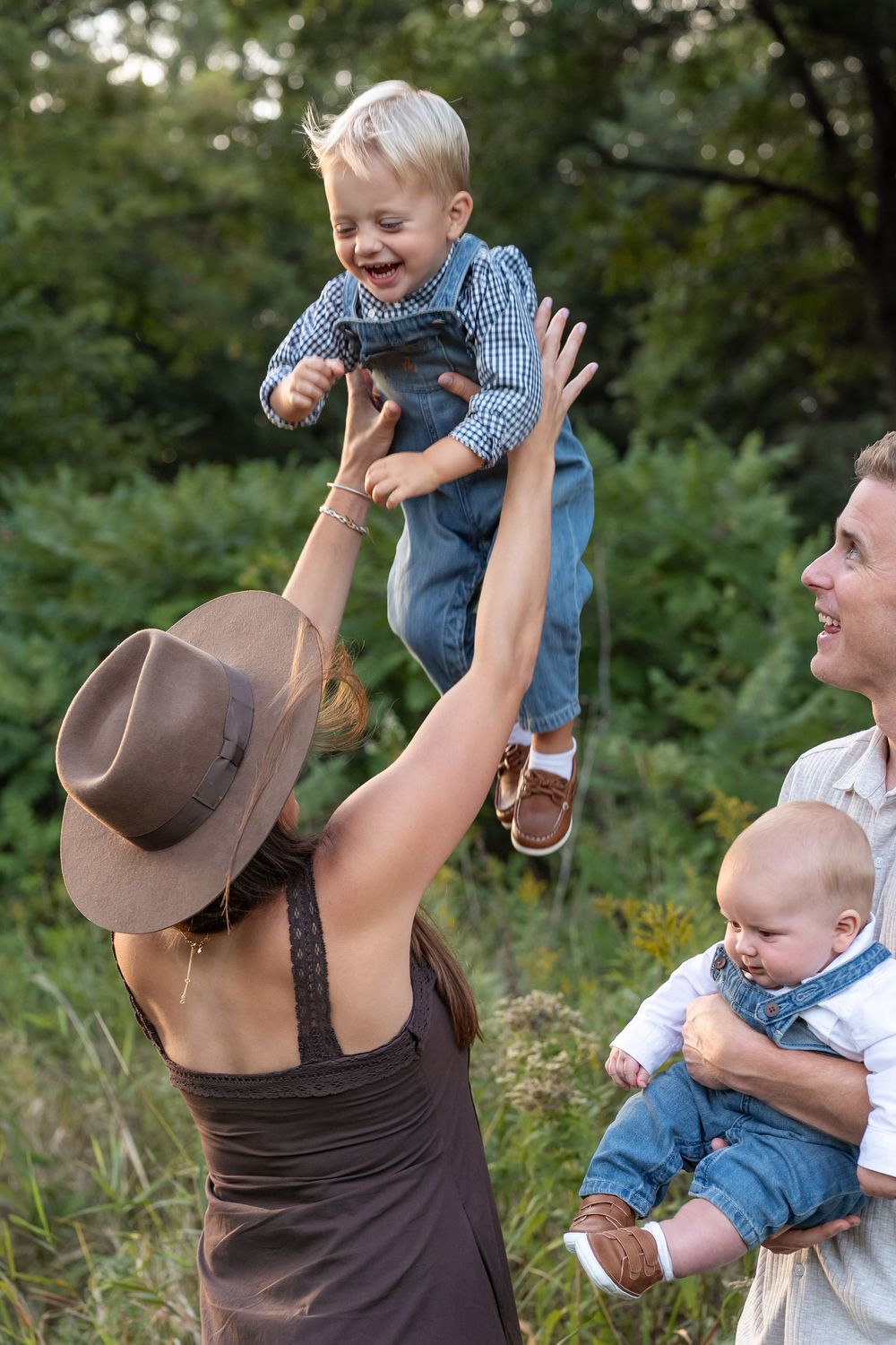 Woman in a brown felt hat lifts a laughing toddler in denim overalls into the air while a smiling man holds a baby in a sunny green field.