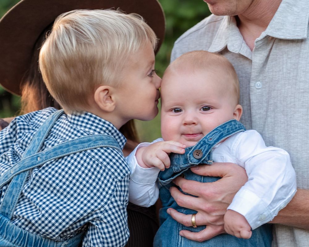 A toddler in a blue gingham shirt and denim overalls kisses the cheek of a smiling baby in denim overalls held by an adult outdoors.