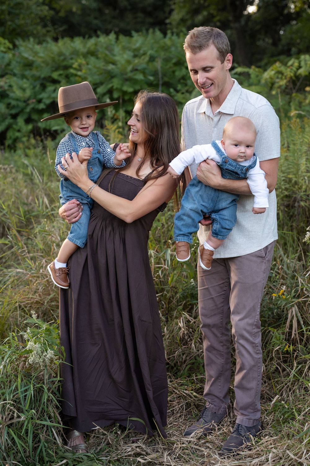 Smiling parents hold two babies, one in a brown hat and denim outfit, in a sunlit grassy field during a family photo session.