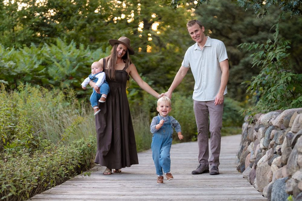Family of four walking on wooden bridge path surrounded by lush greenery, parents holding hands with toddler son.