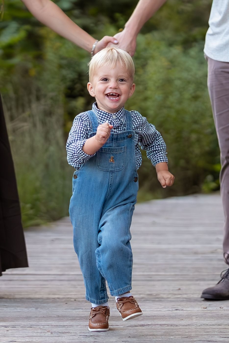 Smiling toddler in denim overalls and plaid shirt walks on wooden path held by adults.