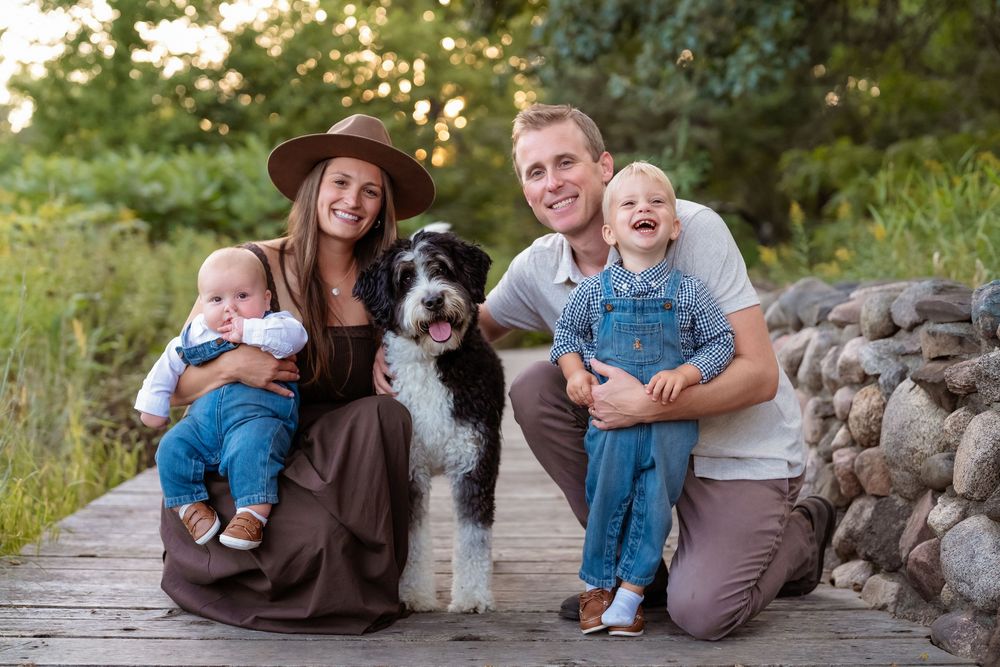 Happy family with two young children and a black-and-white dog posing outdoors on a wooden deck near a stone wall.