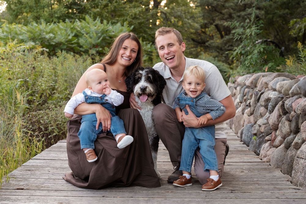 Happy family with two toddlers and a large dog posing on a wooden bridge surrounded by lush greenery.
