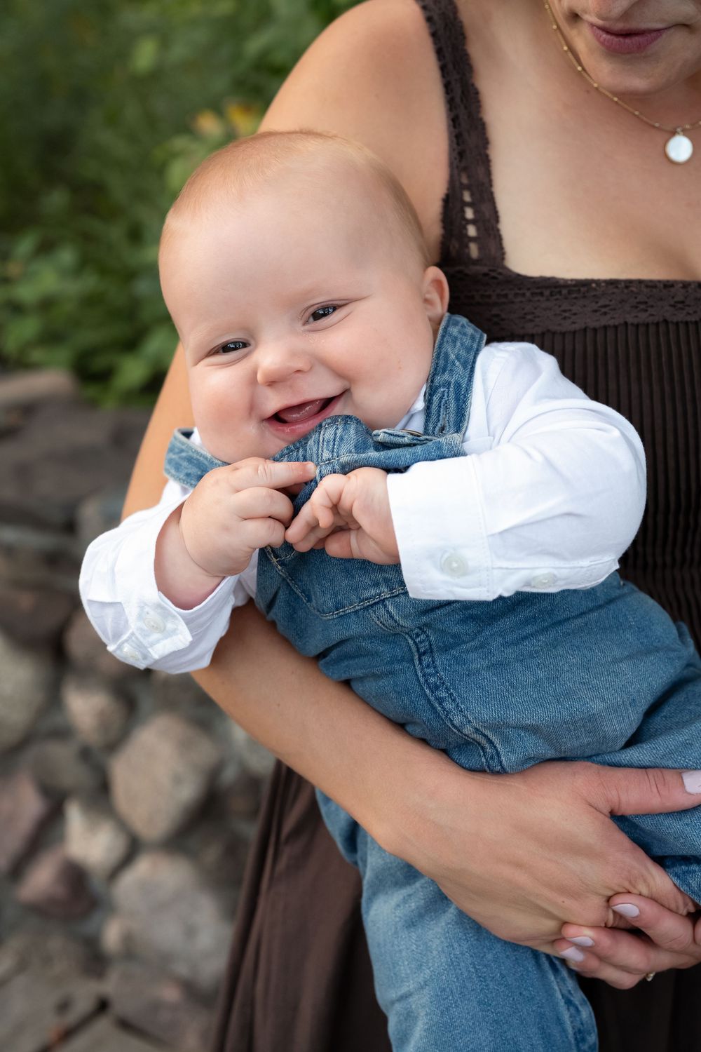 Smiling baby in denim overalls and a white shirt is held in the arms of a woman near a stone wall and green foliage.