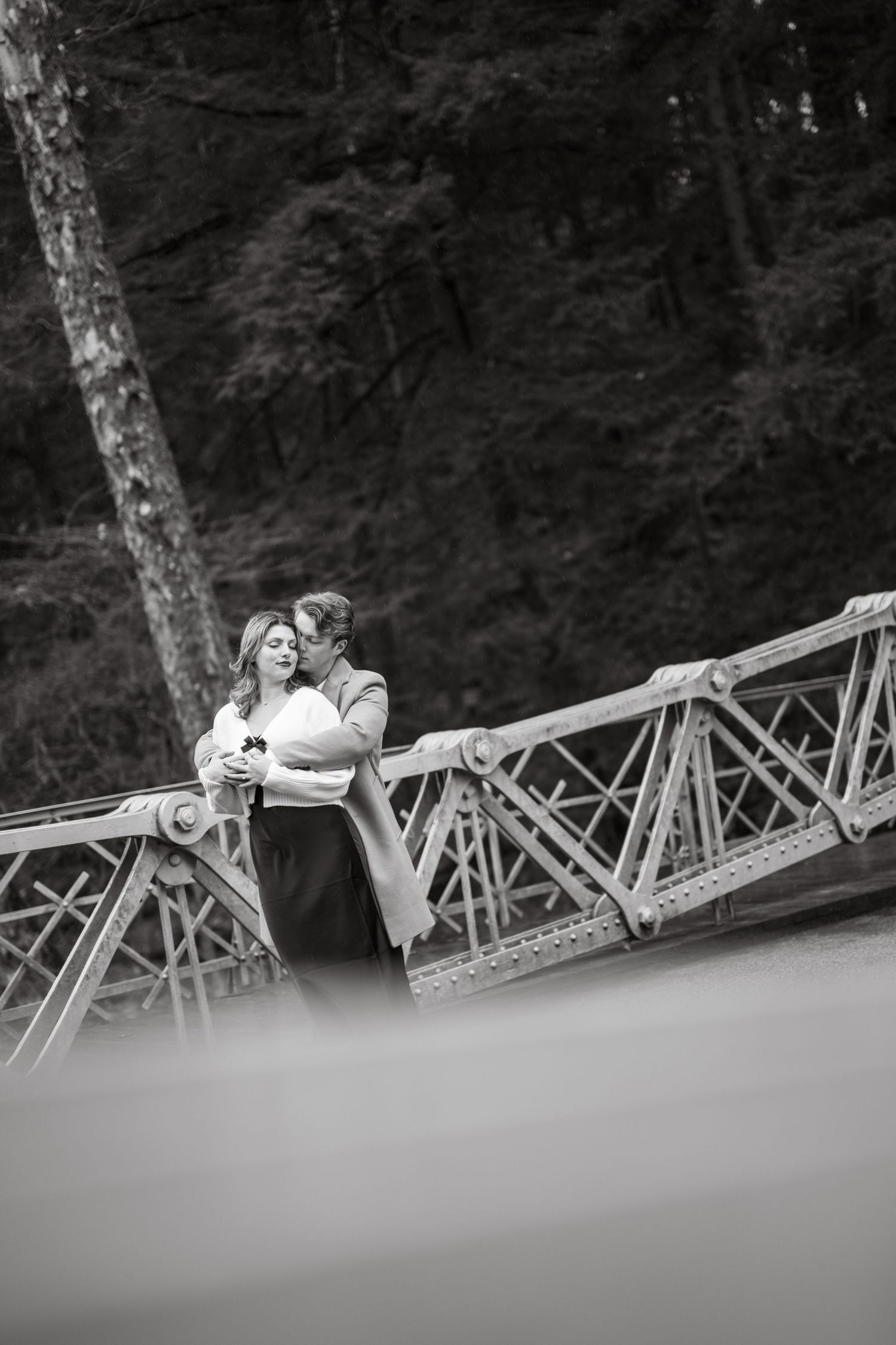 A romantic black and white portrait of a couple embracing on a rustic bridge with trees in the background.