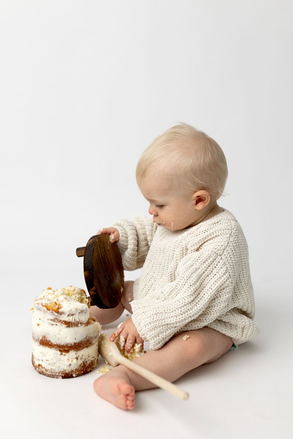 Blonde toddler in cream knit sweater smashing first birthday cake on white background.