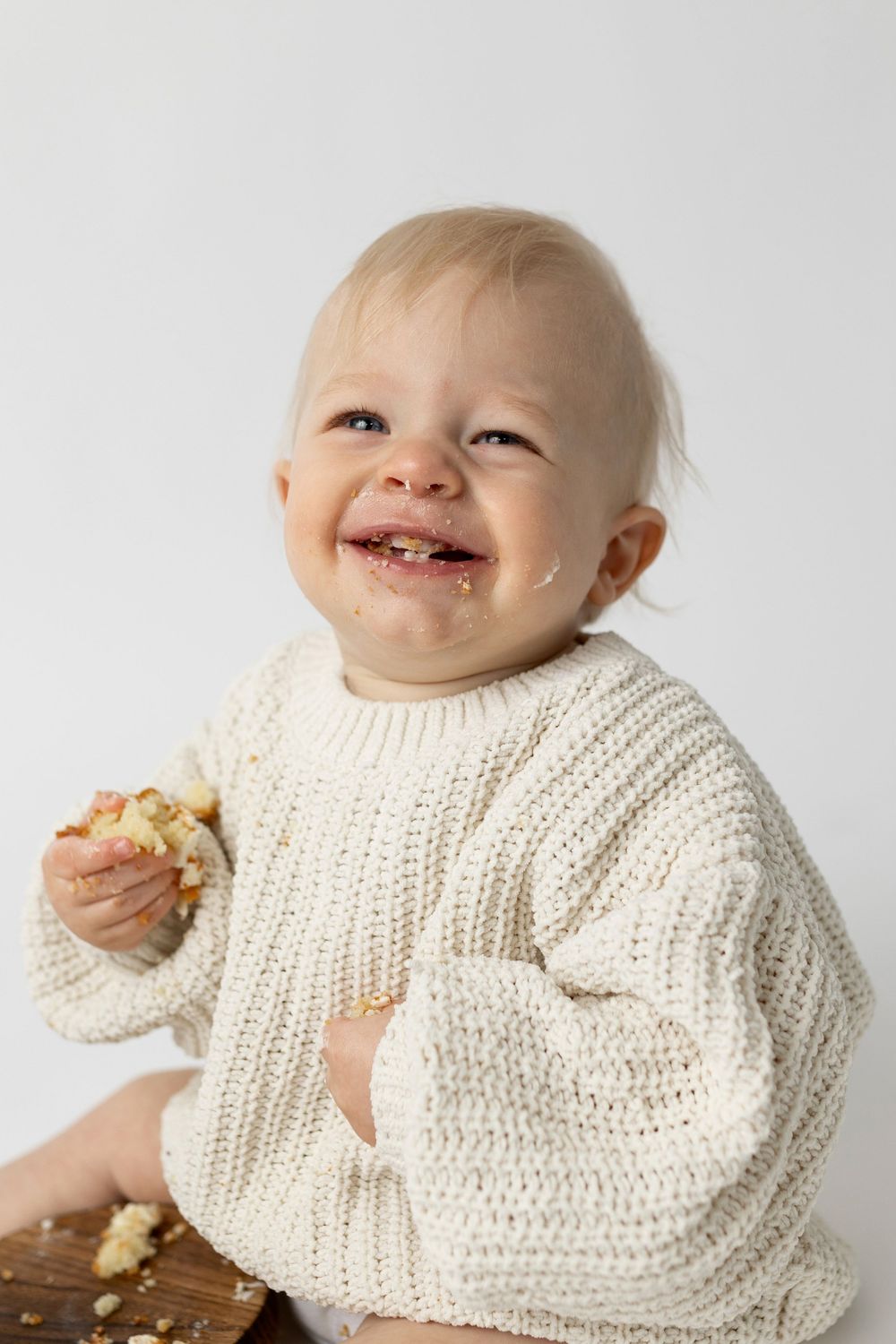 Happy baby in cream knit sweater smiling with food on face, holding a piece of cake during first birthday smash.