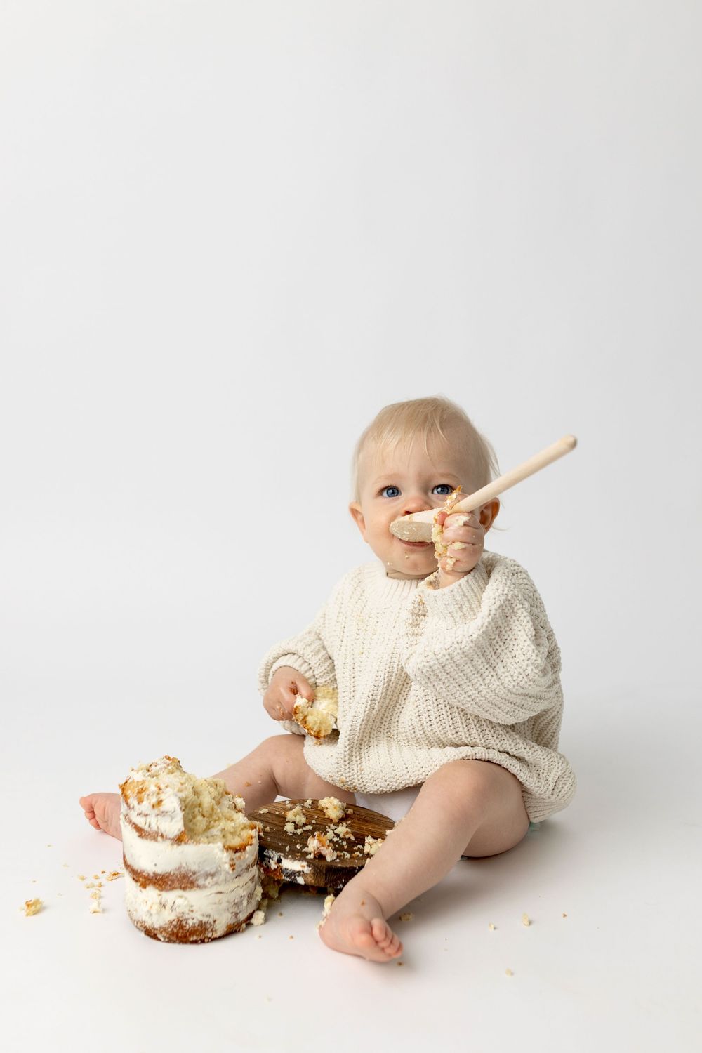 Baby in cream knit romper smashing first birthday cake, licking frosting off a wooden spoon on white background.