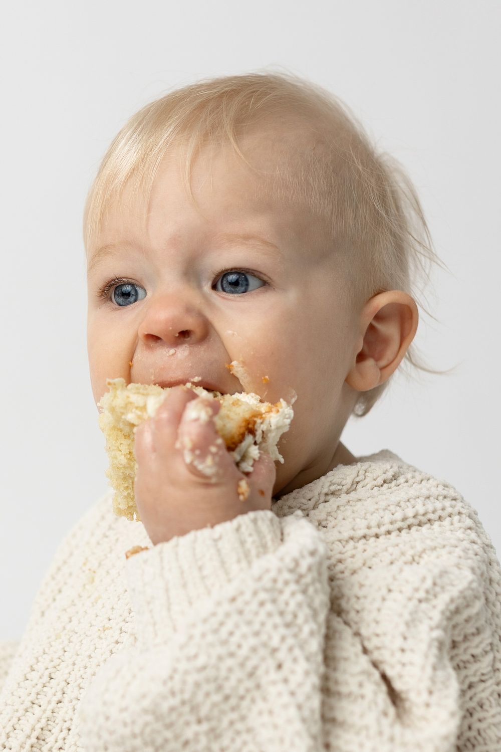 Blonde blue-eyed baby in cream knit sweater messily eating cake with frosting on face and hands.