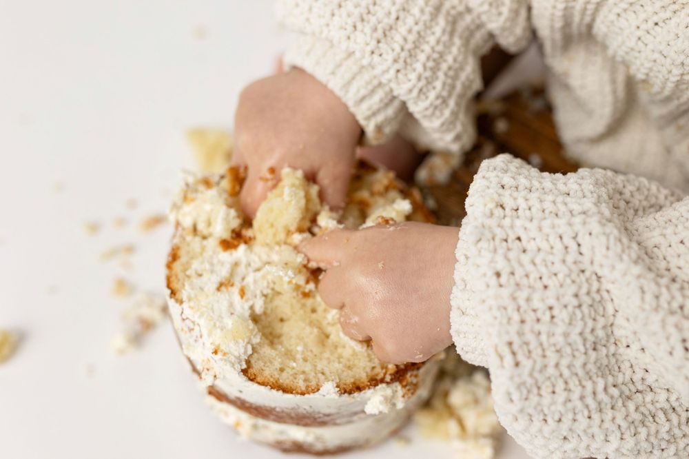 Child's hands breaking apart a white frosted cake, with crumbs scattered on a white surface.
