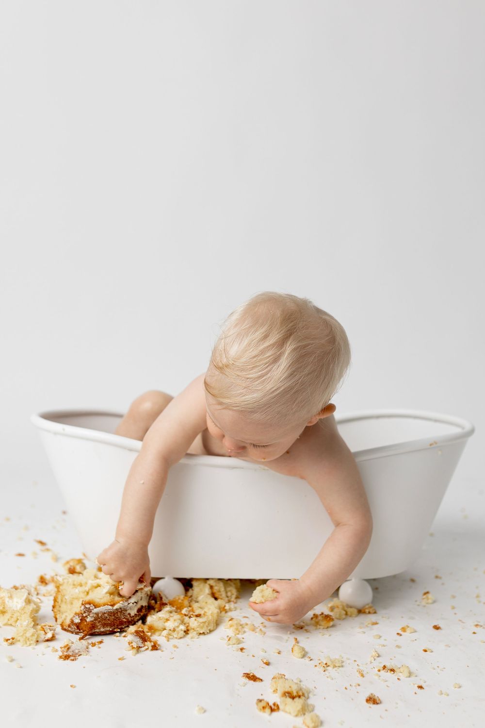 Blonde toddler reaching for smashed cake beside a white mini bathtub during a cake smash session.